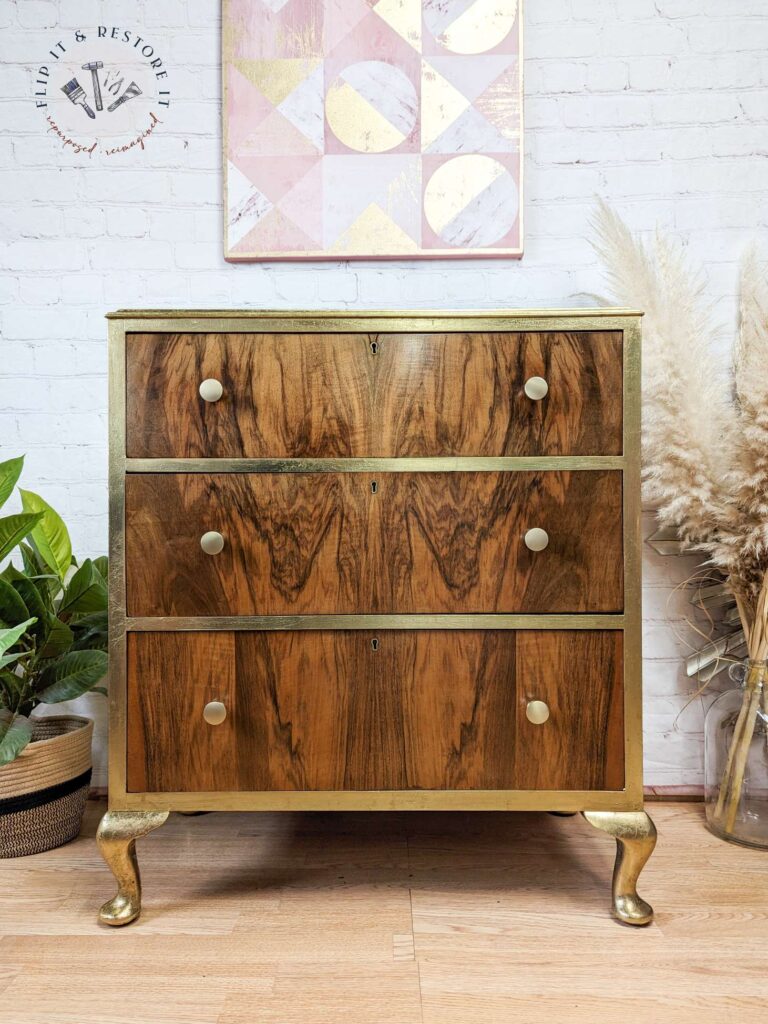 A vintage wooden chest of drawers with three drawers, brass knobs, and gold-painted legs is placed against a white brick wall. Above it hangs a geometric art piece with pastel colors. Plants and dried foliage adorn the surroundings, enhancing the rustic decor.