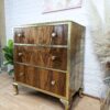 A vintage wooden dresser with three drawers featuring brass fittings and legs stands in a room with a white brick wall. The dresser has a glossy finish and round brass knobs on each drawer. Decorative pampas grass in a vase and green plants adorn the space.