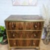 A wooden dresser with three drawers featuring brass handles and edges, placed on a wooden floor. The dresser stands against a white brick wall, adorned with a geometric pattern artwork above. Plants and pampas grass decor surround the furniture.