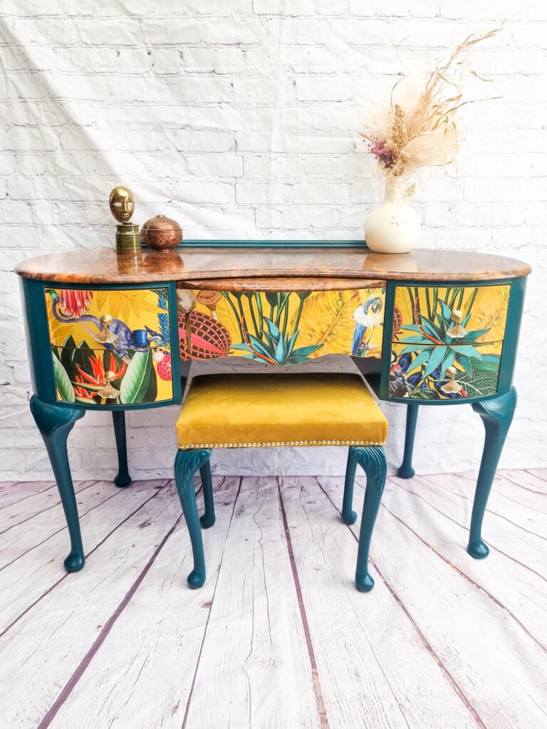 The Queen Anne Painted Burr Walnut Bedroom Set, comprising a dressing table/vanity and a stool, showcases vibrant, tropical floral and bird-themed panels. The vanity desk boasts teal legs supporting a wooden top adorned with a white vase of dried flowers and a small sculpture. Complementing the desk is a stool featuring an inviting mustard-yellow cushion.