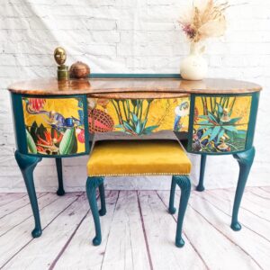 The Queen Anne Painted Burr Walnut Bedroom Set, comprising a dressing table/vanity and a stool, showcases vibrant, tropical floral and bird-themed panels. The vanity desk boasts teal legs supporting a wooden top adorned with a white vase of dried flowers and a small sculpture. Complementing the desk is a stool featuring an inviting mustard-yellow cushion.