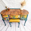 The Queen Anne Painted Burr Walnut Bedroom Set comprises a vintage wooden vanity table with curved edges and a marble-like top. The front showcases vibrant painted plant motifs, and an upholstered yellow velvet stool is nestled underneath. The tabletop is adorned with various decorative items, including a vase filled with dried flowers.