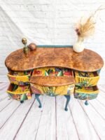 A Queen Anne Painted Burr Walnut Dressing Table from the Bedroom Set, featuring six open, floral-patterned drawers. The top is adorned with two small sculptures and a vase with dried flowers. The legs of the table are painted blue-green. It is set against a backdrop of a white brick wall and light wood flooring.