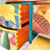 A close-up of the Queen Anne Painted Burr Walnut Bedroom Set's dressing table reveals a colorful, eclectic piece featuring two drawers partially open. The drawers boast a vibrant, whimsical design with images of nature, including plants and mushrooms, intertwined with abstract elements. The dresser is complemented by a teal frame and wooden top.