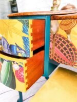 A close-up of the Queen Anne Painted Burr Walnut Bedroom Set's dressing table reveals a colorful, eclectic piece featuring two drawers partially open. The drawers boast a vibrant, whimsical design with images of nature, including plants and mushrooms, intertwined with abstract elements. The dresser is complemented by a teal frame and wooden top.