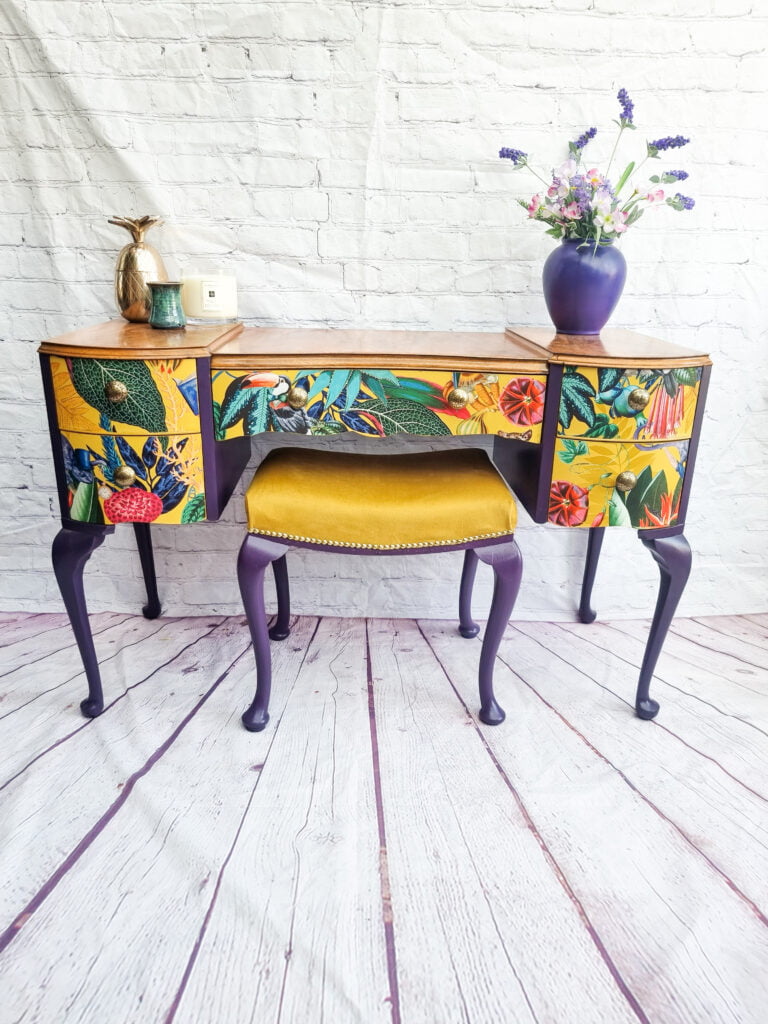 The Queen Anne Painted Walnut Bedroom Set features a dressing table adorned with floral and botanical motifs on its drawers, complemented by elegant purple legs. The set includes an accompanying upholstered stool in a cheerful yellow hue. A vase filled with flowers and a decorative pineapple sit atop the table, all set against a backdrop of white brick walls and a striking purple wooden floor.