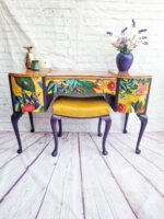 The Queen Anne Painted Walnut Bedroom Set features a dressing table adorned with floral and botanical motifs on its drawers, complemented by elegant purple legs. The set includes an accompanying upholstered stool in a cheerful yellow hue. A vase filled with flowers and a decorative pineapple sit atop the table, all set against a backdrop of white brick walls and a striking purple wooden floor.