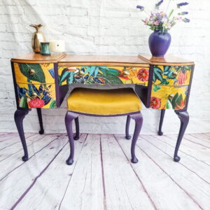 The Queen Anne Painted Walnut Bedroom Set features a dressing table adorned with floral and botanical motifs on its drawers, complemented by elegant purple legs. The set includes an accompanying upholstered stool in a cheerful yellow hue. A vase filled with flowers and a decorative pineapple sit atop the table, all set against a backdrop of white brick walls and a striking purple wooden floor.