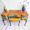 The Queen Anne Painted Walnut Bedroom Set, featuring a dressing table/vanity and stool, stands against a white brick wall and light wooden floor. The vanity boasts colorful, tropical-themed drawer fronts complemented by purple-painted legs. Atop the vanity are candles, a small pineapple decoration, and a purple vase holding an arrangement of flowers.