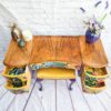 The Queen Anne Painted Walnut Bedroom Set, featuring a dressing table/vanity and a yellow stool, showcases a vintage wooden desk with an intricate wood grain top. The sides of the drawers are adorned with colorful floral patterns, which continue into the partially open drawer interiors. A vase with flowers, a candle, and other decorative items grace the top of the table.