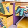 A colorful dresser from the Queen Anne Painted Walnut Bedroom Set showcases open drawers revealing a bright interior. The fronts of the drawers are adorned with images of a toucan, cactus, and foliage, complemented by purple sides. The top surface holds a candle and a decorative piece, with part of a yellow chair visible nearby.