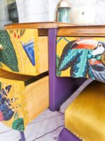 A colorful dresser from the Queen Anne Painted Walnut Bedroom Set showcases open drawers revealing a bright interior. The fronts of the drawers are adorned with images of a toucan, cactus, and foliage, complemented by purple sides. The top surface holds a candle and a decorative piece, with part of a yellow chair visible nearby.