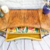 Top view of a Queen Anne Painted Walnut Dressing Table against a white brick wall. The table features a textured wood surface, with an open drawer lined with bright botanical print paper. A candle, green pot, and purple vase with flowers decorate the top of the dressing table.