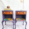 A Queen Anne Painted Walnut Bedroom Set dressing table with ornate legs is displayed against a white brick wall. The table features a wooden drawer and a colorful, vibrant bottom shelf. On top of the table, there are stacked books and a small round container on one side, while the other side showcases a lamp with a round bulb.