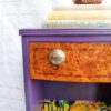 The Queen Anne Painted Walnut Bedroom Set - Dressing Table/Vanity and Stool features a small, vividly painted dressing table with a top drawer boasting a wood grain finish and a round, textured knob. The tabletop holds several books and a brass pot, while the shelf below displays colorful items that contribute to an eclectic decor. The setup is beautifully complemented by the white brick wall in the background.