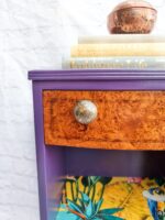 The Queen Anne Painted Walnut Bedroom Set - Dressing Table/Vanity and Stool features a small, vividly painted dressing table with a top drawer boasting a wood grain finish and a round, textured knob. The tabletop holds several books and a brass pot, while the shelf below displays colorful items that contribute to an eclectic decor. The setup is beautifully complemented by the white brick wall in the background.