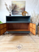 The Green and Gold Painted MCM Nathan Cabinet/Media Unit/Entertainment Sideboard stands with its doors open, revealing an empty wooden interior. It boasts a sleek dark green external paint, complemented by gold-tipped legs and a decorative display shelf on top. The scene is set against a backdrop of a white brick wall, potted plants, and a landscape painting.