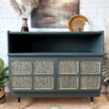 The Green and Gold Painted MCM Nathan Cabinet/Media Unit/Entertainment Sideboard showcases a stunning dark finish, complemented by intricate gold accents on its cabinet doors. It stands elegantly on tapered legs with gold tips. The top is adorned with stylish decor items like dried plants, a framed picture, and a ceramic bird. Above the sideboard hangs a charming rustic painting.