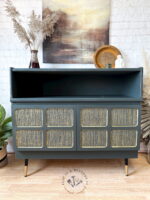 The Green and Gold Painted MCM Nathan Cabinet/Media Unit/Entertainment Sideboard showcases a stunning dark finish, complemented by intricate gold accents on its cabinet doors. It stands elegantly on tapered legs with gold tips. The top is adorned with stylish decor items like dried plants, a framed picture, and a ceramic bird. Above the sideboard hangs a charming rustic painting.