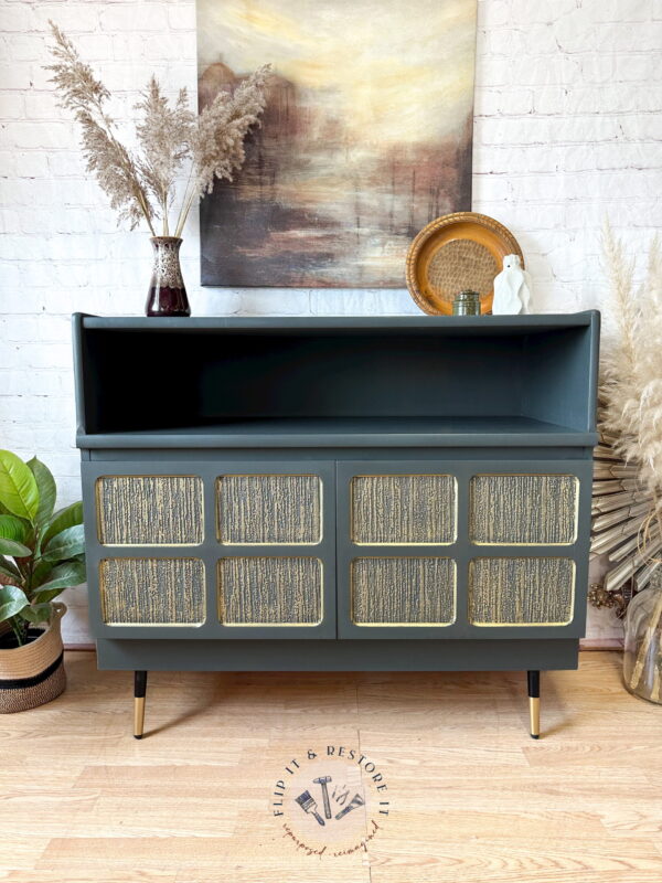 A refurbished Green and Gold Painted MCM Nathan Cabinet/Media Unit/Entertainment Sideboard stands against a white brick wall. Behind it is a landscape painting, while to the left, there is a plant. On top of the sideboard to the right are a wicker basket and pampas grass in a vase.