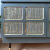 A close-up of the Green and Gold Painted MCM Nathan Cabinet/Media Unit/Entertainment Sideboard, showcasing its modern green wooden design with textured gold inlays on the doors. The cabinet stands on sleek tapered legs featuring black and gold accents. The background includes a light wall and wooden floor. A logo with the text "Refurb & Restore Ltd." is visible at the bottom.