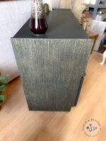 A close-up of the Green and Gold Painted MCM Nathan Cabinet/Media Unit/Entertainment Sideboard with a textured surface painted in dark green and gold vertical streaks sits on a hardwood floor. A maroon vase with a textured finish is placed on top of the cabinet. Decor items and a plant are visible in the background.