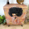 A Hand-painted Cabinet adorned with artwork of a woman with an afro on its doors, featuring a light brown finish, gold handles, and metal legs. Surrounding it are houseplants and decorative items like a small vase and a wooden bowl.