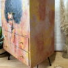 A hand-painted linen cupboard with two drawers is finished in warm tones and showcases an artistic portrait of a woman with curly black hair on the left side. The tallboy cupboard stands on sleek black metal legs and is set against a white brick wall, with dried grasses in a vase visible on the right.