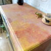 A close-up of a decorated Hand-painted cabinet with a distressed pink, orange, and yellow finish. On top are a ceramic vase with intricate patterns, a small gold elephant figurine, and a wooden box with a black design. A logo reading "Flip it & Restore it" is on the wall.