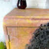 Close-up of a hand-painted Tallboy cabinet, featuring a textured finish in vibrant yellow and pink tones. A vase with intricate brown detailing sits on top. The background showcases a white brick wall. In the upper left corner, the "Flip It + Restore It" logo is partially visible.