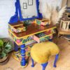 The Vintage Victorian Mahogany Duchess Dressing Table Painted Bedroom Set includes a beautifully painted vanity in blue and yellow with floral patterns, featuring several small drawers. Accompanied by an upholstered stool with a yellow velvet seat, the set is elegantly positioned on a wooden floor, surrounded by potted plants and decor.