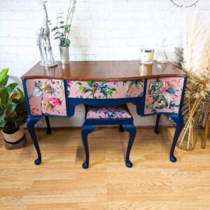 A Queen Anne Painted Mahogany Bedroom Set, featuring a vanity and stool with blue finish and tropical bird and floral patterns on the drawer fronts and cushion, stands against a white brick wall. The tabletop is adorned with a potted plant, small sculpture, candle, and framed photo. A logo reads "Flip It & Restore It.