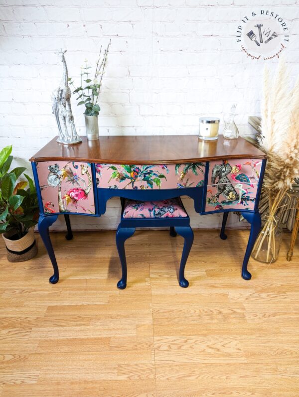 A Queen Anne Painted Mahogany Bedroom Set, featuring a vanity and stool with blue finish and tropical bird and floral patterns on the drawer fronts and cushion, stands against a white brick wall. The tabletop is adorned with a potted plant, small sculpture, candle, and framed photo. A logo reads "Flip It & Restore It.