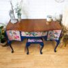 A Queen Anne Painted Mahogany Bedroom Set, featuring a dressing table/vanity adorned with floral and bird motifs on its drawers and sides, painted in dark blue, stands against a white brick wall. On top of the vanity rests an assortment of items including a giraffe statue, two vases, and a candle. A matching stool with a floral seat is tucked beneath it.