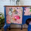 The Queen Anne Painted Mahogany Bedroom Set dressing table features a vibrantly painted design with wildlife illustrations, including a koala, flamingo, giraffe, and parrot on the drawer fronts. The primarily blue piece has a pink background for the animals and is topped with decorative giraffe figurines and a potted plant.