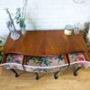 A Queen Anne Painted Mahogany Dressing Table, from a Bedroom Set, viewed from above. Two partially open drawers reveal colorful, floral-themed linings with bird illustrations. The table's top has decorative items including a giraffe sculpture, a vase with greenery, and a candle.