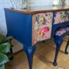 A Queen Anne Painted Mahogany Dressing Table with deep blue legs and sides, featuring drawers adorned with colorful animal artwork including birds and mammals, stands in front of a brick wall. The wooden tabletop is complemented by a plant, a candle, and silver animal figurines.