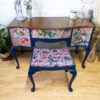 The Queen Anne Painted Mahogany Bedroom Set includes a dressing table/vanity and stool, both boasting blue legs. The stool and drawer fronts are upholstered with vibrant floral and bird-themed fabric. The vanity is adorned with plants, a candle, and a clock, creating an elegant scene against a white brick wall.