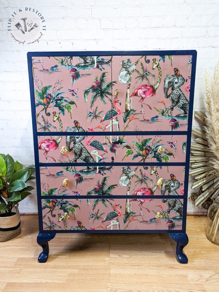 Presenting the Vintage Tallboy Chest Of Drawers Linen Cupboard: a decorative piece featuring a vibrant tropical bird design with flamingos, parrots, and peacocks set against a pink background. The dresser boasts dark blue edges and legs. It's showcased on a wooden floor next to a potted plant and dried pampas grass.