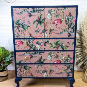 Presenting the Vintage Tallboy Chest Of Drawers Linen Cupboard: a decorative piece featuring a vibrant tropical bird design with flamingos, parrots, and peacocks set against a pink background. The dresser boasts dark blue edges and legs. It's showcased on a wooden floor next to a potted plant and dried pampas grass.