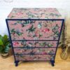 The Vintage Tallboy Chest Of Drawers Linen Cupboard has been refurbished with a vibrant tropical print showcasing flamingos, palm trees, and various exotic birds on a pink background. The edges are painted navy blue, and the chest stands on four small legs, accompanied by a natural wooden floor and decorative plants beside it.