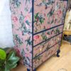 The Vintage Tallboy Chest Of Drawers Linen Cupboard offers a striking combination of vintage charm and bold design. This dresser stands on dark blue legs and is embellished with a vibrant animal print, showcasing multiple drawers decorated with illustrations of various birds and wildlife against a pink background. Green plants and decor items nearby enhance its picturesque appeal.