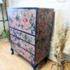 The Vintage Tallboy Chest Of Drawers Linen Cupboard is a delightful four-drawer dresser adorned with a whimsical pink pattern that includes floral designs and charming animals like birds, set amidst lush foliage. The dresser stands on dark blue legs, perfectly complementing its playful design. It is pictured against a white brick wall with a vase of tan pampas grass beside it and a spotlight partially visible on the left.