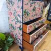 A vintage wooden dresser, known as the "Vintage Tallboy Chest Of Drawers Linen Cupboard," is adorned with a pink tropical-themed wallpaper featuring palm trees, flamingos, and other exotic wildlife. This charming piece boasts three open drawers that reveal the natural wood inside. Green plants surround the dresser, with a logo displayed on the floor nearby.