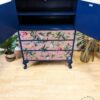 A refurbished dark blue Vintage Tallboy Chest Of Drawers Linen Cupboard with the doors open, revealing two shelves. The cupboard features intricate botanical and bird designs on the drawer fronts, painted in various colors. It stands on a light wood floor, with plants and dried grasses beside it.