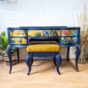 The Vintage French Style Painted Bedroom Set - Dressing Table/Vanity and Stool features a vibrant blue vintage vanity desk with three ornately painted drawers adorned with colorful nature and bird motifs. The desk boasts elegant curved legs and is complemented by a matching blue stool upholstered in mustard yellow fabric. The setup also includes a backdrop of a white brick wall and pampas grass decor.