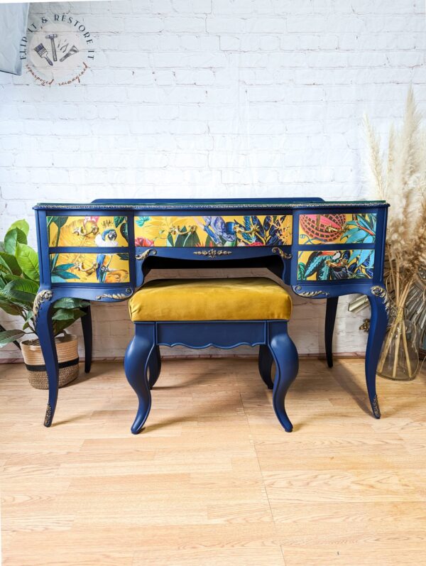 The Vintage French Style Painted Bedroom Set - Dressing Table/Vanity and Stool features a vibrant blue vintage vanity desk with three ornately painted drawers adorned with colorful nature and bird motifs. The desk boasts elegant curved legs and is complemented by a matching blue stool upholstered in mustard yellow fabric. The setup also includes a backdrop of a white brick wall and pampas grass decor.