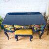 The Vintage French Style Painted Bedroom Set features a stunning ornate blue dressing table with a glossy finish, showcasing colorful, intricate designs on the front panels. Accompanying the set is a matching blue and yellow cushioned stool that fits neatly beneath the table. This elegant setup is displayed on a wooden floor against a white brick wall, accented by nearby plants.