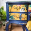 A close-up of the vividly painted dressing table from the Vintage French Style Painted Bedroom Set, featuring two drawers. The table is mainly blue with intricate gold details, and the drawer fronts showcase colorful illustrations of parrots and tropical leaves. A plant in the background and a part of a stool are also partially visible.