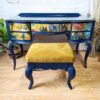 The Vintage French Style Painted Bedroom Set - Dressing Table/Vanity and Stool showcases a vividly painted blue vanity desk adorned with intricate, colorful floral and bird designs on the drawers. The set includes a matching cushioned stool upholstered in vibrant yellow fabric. It is elegantly displayed on a light wooden floor against a white brick wall.